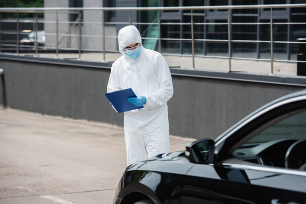 Trabajador médico en traje de materiales peligrosos y gafas de escribir en el portapapeles cerca de coche borroso  - Foto, Imagen