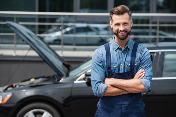 Positive mechanic in overalls looking at camera near blurred car  - Foto, Bild