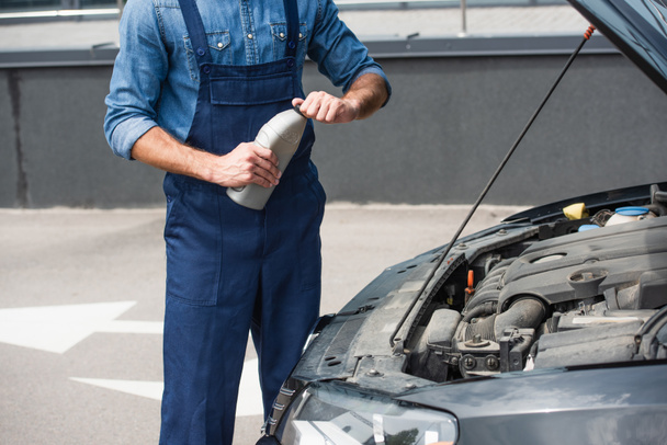 Oříznutý pohled mechanika držícího láhev motorového oleje v blízkosti auta s otevřenou kapotou  - Fotografie, Obrázek