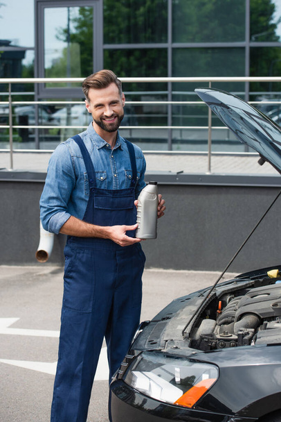 Smiling mechanic holding bottle of motor oil near car outdoors  - Foto, imagen