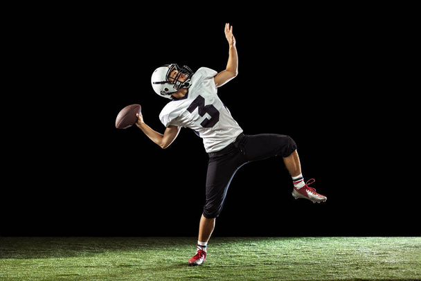 One professional American football player playing football isolated on dark studio background with green grass flooring. Concept of sport, competition - Foto, afbeelding