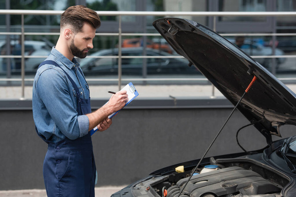 Side view of mechanic in overalls writing on clipboard near car outdoors  - 写真・画像
