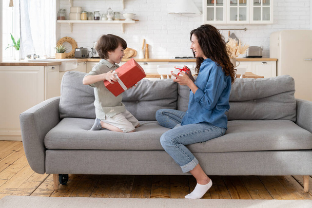 Mom and son celebrate birthday together: happy mum and excited son sit on couch hold gifts smiling - Fotó, kép