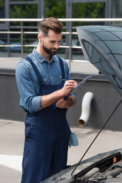 Mechanic in overalls writing on clipboard near car with open hood  - Фото, изображение