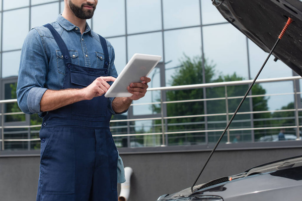 Cropped view of mechanic in overalls using digital tablet near car outdoors  - Foto, afbeelding