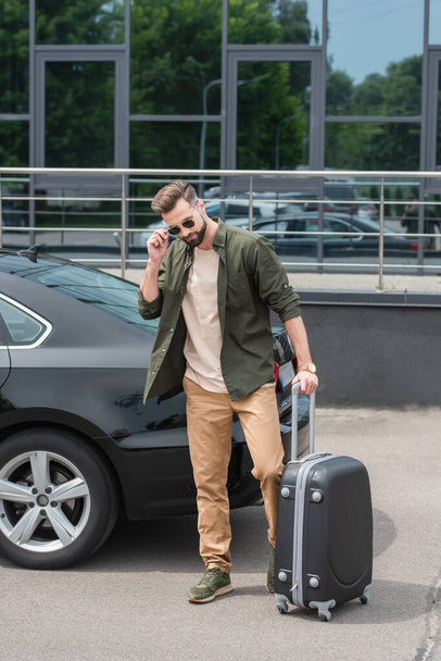Tourist in sunglasses holding suitcase near car outdoors  - Foto, imagen