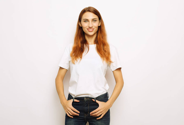 Stylish young woman with long red hair wearing a white t-shirt and jeans posing on a white background. Fashion concept. - Photo, image