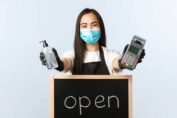 Small business, covid-19 pandemic, virus and contactless orders concept. Friendly pleasant smiling asian waitress, female barista giving visitors payment terminal and hand sanitizer, show open sign - Photo, Image
