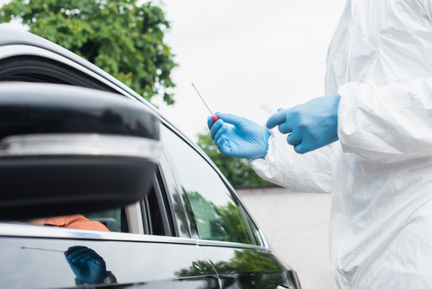 Widok pracownika medycznego w kombinezonie ochronnym przeprowadzającego test pcr w pobliżu kierowcy w nieostrym samochodzie - Zdjęcie, obraz