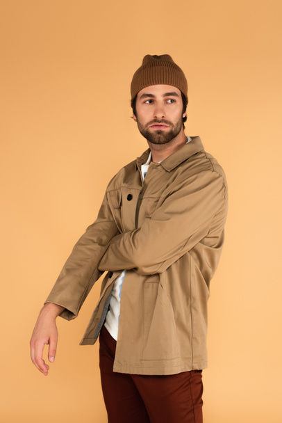 young man in jacket and brown beanie looking away while standing isolated on beige - Foto, immagini