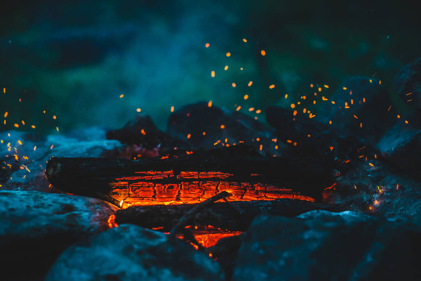 Élénk parázsló tűzifa égett a tűzben közelről. Légköri háttér narancssárga lánggal a tábortűz és a kék füst. A tábortűz teljes képét. Ragyogó parázs a levegőben. Fényes szikrák bokeh-ban - Fotó, kép
