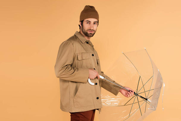 man in stylish autumn outfit looking at camera while folding umbrella isolated on beige - Foto, Bild