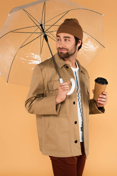 positive man with paper cup looking back under transparent umbrella isolated on beige - Foto, Imagen
