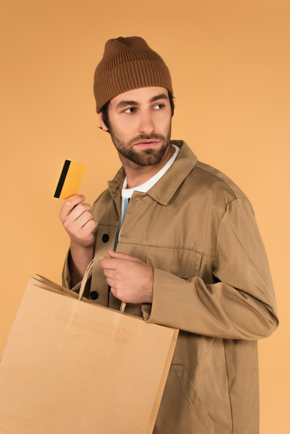 молодой человек в шапочке и куртке, держащий сумки с покупками и кредитные карты изолированы на бежевый - Фото, изображение