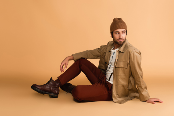 trendy man in stylish autumn outfit looking away while sitting on beige - Valokuva, kuva