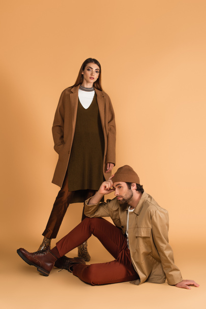 young and stylish woman looking at camera near trendy man sitting on beige background - Foto, immagini