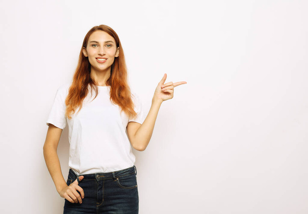Smiling young red-haired woman isolated on gray studio background pointing with finger at blank copy space aside, happy positive female show at vacant advertising spot - Fotografie, Obrázek