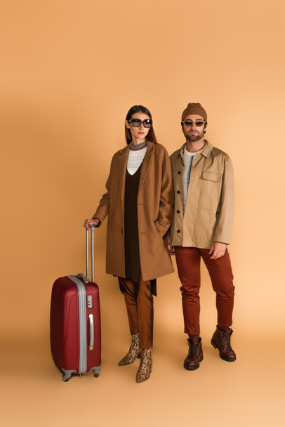 young fashionable couple in sunglasses near suitcase on beige background - Φωτογραφία, εικόνα