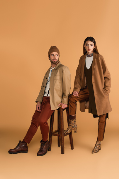 woman with hand in pocket of stylish coat near trendy man sitting on high stool on beige background - Foto, immagini