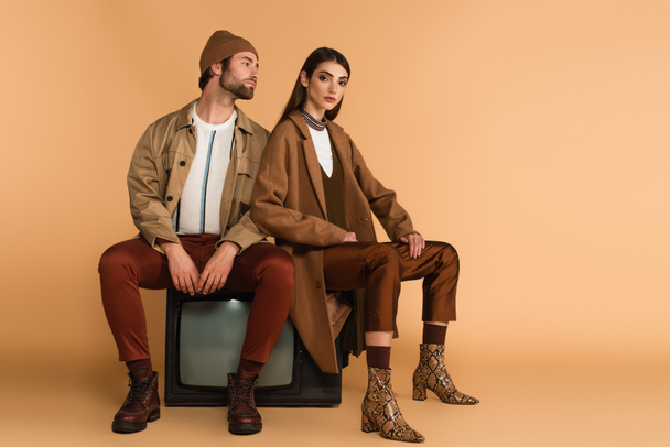 young couple in fashionable autumn outfit sitting on vintage tv set on beige background - Foto, Imagem