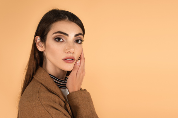 young brunette woman touching face while looking at camera isolated on beige - Photo, Image