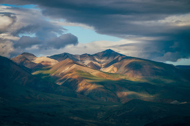 Φοβερό ορεινό τοπίο με μεγάλα βουνά και μπλε κάθαρση στο συννεφιασμένο ουρανό σε συννεφιασμένο καιρό. Μπλε φεγγίτης σε συννεφιασμένο ουρανό πάνω από τεράστια πολύχρωμα βουνά στο φως του ήλιου. Πολύχρωμο ορεινό τοπίο - Φωτογραφία, εικόνα