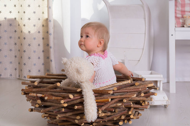A little one-year-old girl in a white and pink dress sits with a soft toy white bear in a nest of wooden sticks in a room with an interior in light colors - Foto, Imagen
