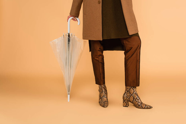 részleges kilátás nő bőrcsizmában és barna nadrág támaszkodik esernyő bézs háttér - Fotó, kép