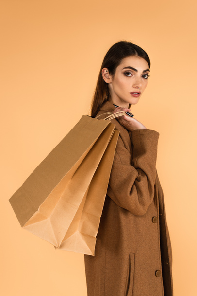 stylish brunette woman with shopping bags looking at camera isolated on beige - Fotó, kép