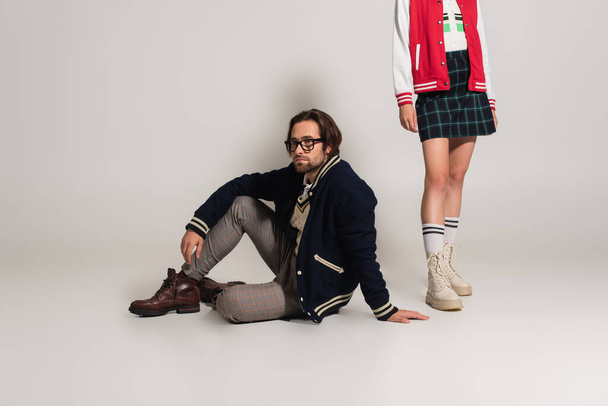 stylish man in blue jacket sitting near man in plaid skirt and boots on grey background - Foto, imagen