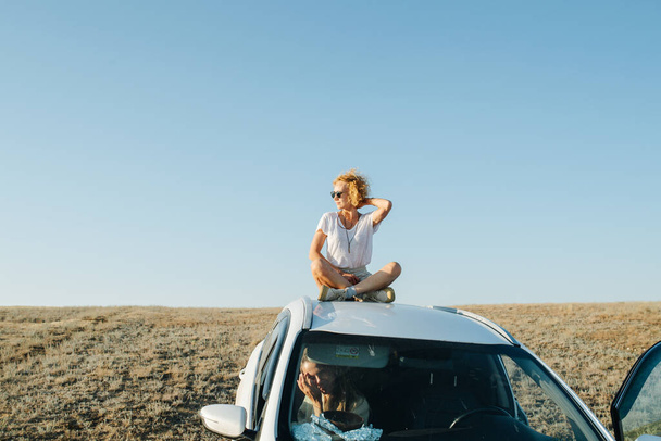 車の屋根の上に座っている楽しい女性は、風と景色の良い草原の草原の景色を楽しんでいます。彼女の友人は車の中だ. - 写真・画像