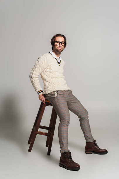 young man in white sweater and pants sitting on high wooden stool on grey background - Фото, зображення