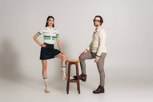 trendy young couple looking at camera while posing near high stool on grey background - Photo, Image