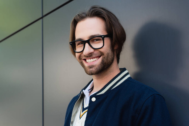 happy young man in stylish eyeglasses standing near grey wall while smiling at camera - Foto, Imagem