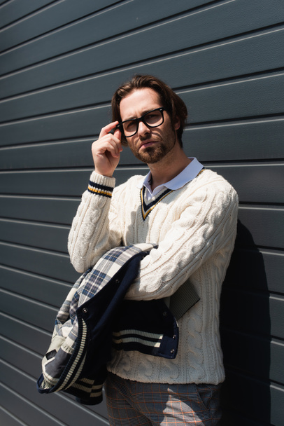 trendy man in white knitted sweater adjusting eyeglasses near grey wall outdoors - Fotó, kép