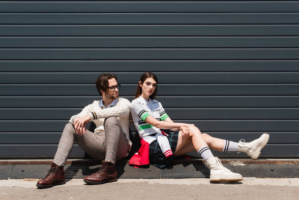 молодая модная пара, сидящая на границе возле серой стены на открытом воздухе - Фото, изображение