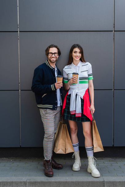 trendy couple with shopping bags and coffee to go smiling near grey wall  - Fotografie, Obrázek