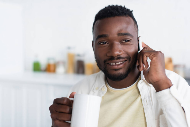 happy african american man holding cup of coffee and talking on cellphone  - Foto, immagini
