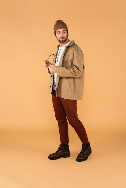full length view of trendy man looking back while holding eyeglasses on beige - Foto, immagini