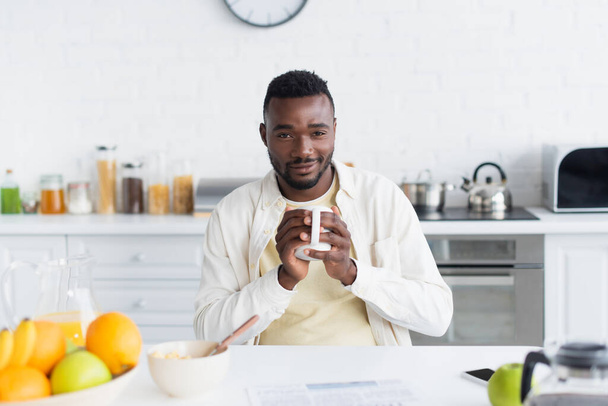 happy african american man holding cup of coffee near breakfast on table - 写真・画像