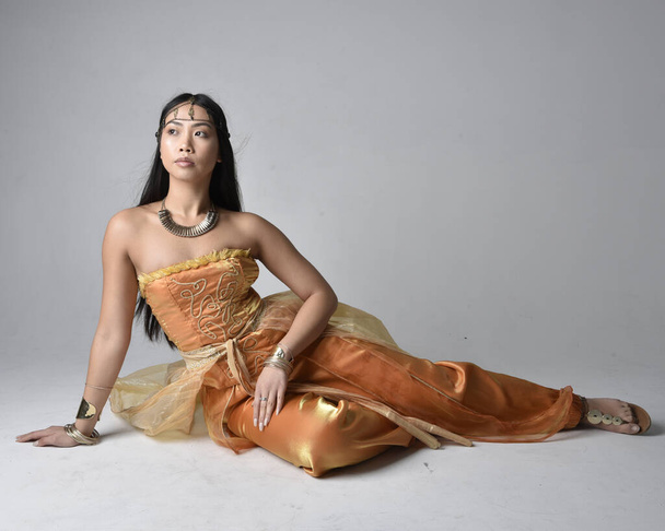 Портрет красивой молодой азиатки в золотых арабских одеждах, как джинн, сидящая поза, изолированная на студийном фоне. - Фото, изображение