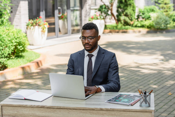joven hombre de negocios afroamericano en gafas usando el ordenador portátil fuera - Foto, imagen