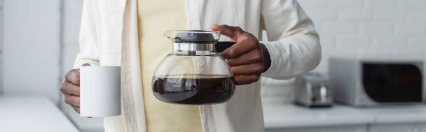 przycięty widok młodego Afroamerykanina trzyma dzbanek do kawy i kubek w kuchni, sztandar - Zdjęcie, obraz
