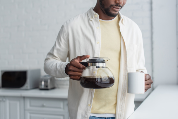 vista recortada de un joven afroamericano sosteniendo cafetera y taza en la cocina - Foto, imagen