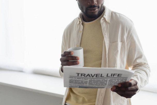cropped view of african american man holding cup of coffee and reading travel life newspaper  - Foto, imagen