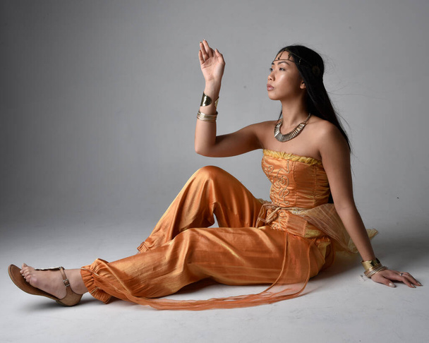 Full length portrait of pretty young asian woman wearing golden Arabian robes like a genie, seated pose, isolated on studio background. - 写真・画像