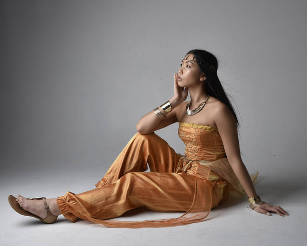 Full length portrait of pretty young asian woman wearing golden Arabian robes like a genie, seated pose, isolated on studio background. - Фото, зображення
