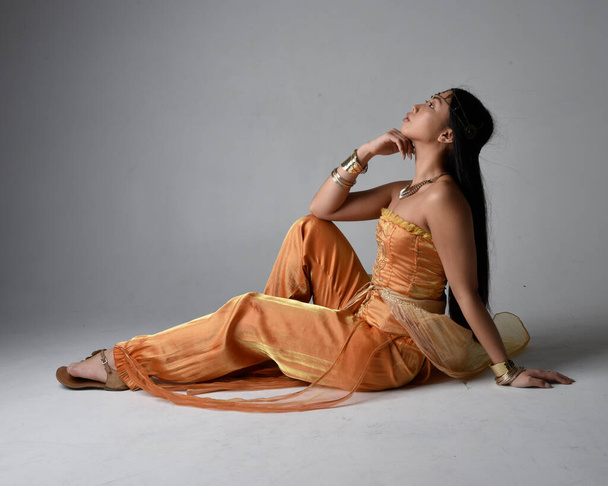 Full length portrait of pretty young asian woman wearing golden Arabian robes like a genie, seated pose, isolated on studio background. - 写真・画像