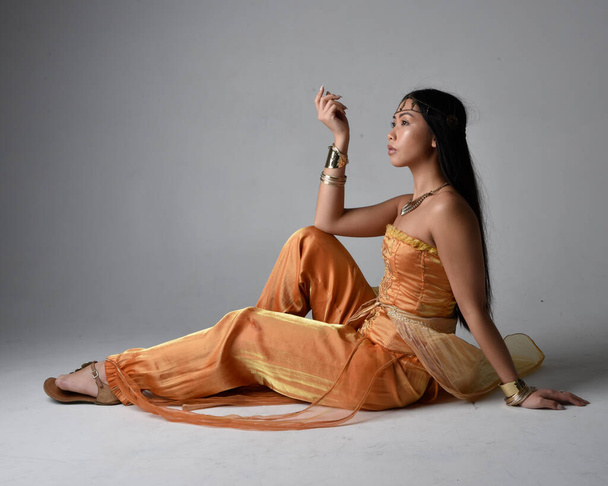 Full length portrait of pretty young asian woman wearing golden Arabian robes like a genie, seated pose, isolated on studio background. - Foto, Bild