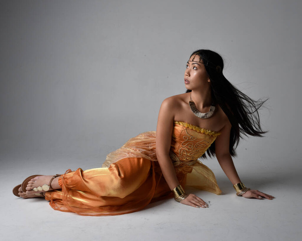 Full length portrait of pretty young asian woman wearing golden Arabian robes like a genie, seated pose, isolated on studio background. - Foto, Imagen